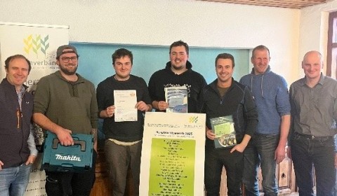 Menschen stehen mit Siegerurkunden nebeneinander in einem Raum. Davor eine Staffelei