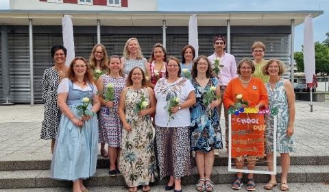 Mehrere Frauen, sowie ien Mann, stehen aufgereiht nebeneinander. Jede hält feierlich eine Rose in der Hand.