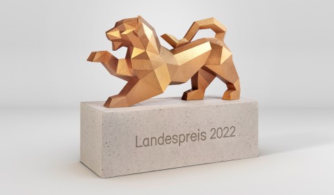 In Form einer Trophäe ist ein güldener stilisierter Wappenlöwe aus dem Landeswappen von Baden-Württemberg auf einem Sockel platziert zu sehen. Auf dem grauen Sockel steht geschrieben: Landespreis 2022.