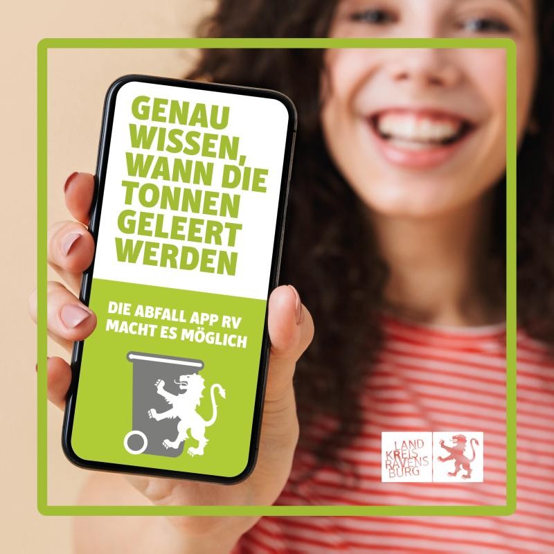 Eine weibliche lächelnde Person hält ein Smartphone mit Bildschirm zur Blickrichtung. Auf dem Bildschirm des Smartphones steht in hellgrüner Schrift auf weißem Grund geschrieben: Genau wissen, wann die Tonnen geleert werden. Darunter steht in weißer Schrift auf hellgrünem Grund: Die Abfall App RV macht es möglich, darunter wiederum das Logo der Abfall App, bestehend aus einer grauen Mülltonne, leicht versetzt davor an deren rechter Seite ein weißer heraldischer Löwe der Welfen, das Wappentier des Landkreises Ravensburg