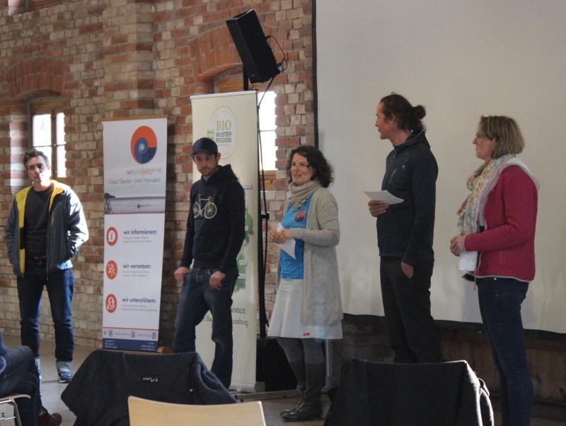 Podiumsgespräch Das Bild zeigt fünf Personen, insgesamt drei Männer und zwei Frauen, die nebeneinander dem Publikum zugewandt vor einer Projektionsleinwand stehen und teilweise Karteikarten in der Hand halten.