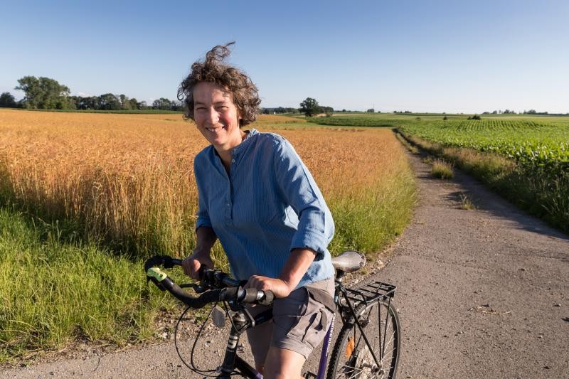 BioGenuss-Radtouren Eine Frau mittleren Alters fährt auf einem Fahrrad eine frühlingshafte oder sommerliche Felderlandschaft unter klarem Himmel entlang.