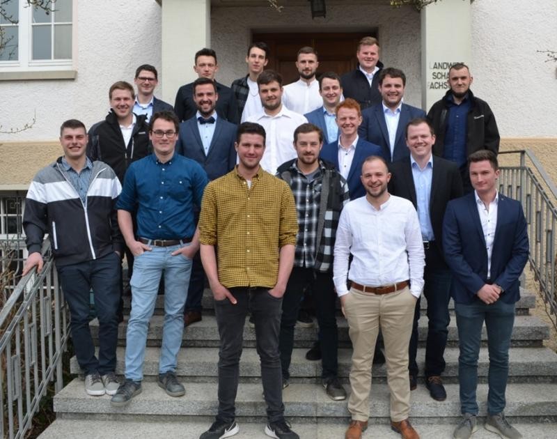 Auf mehreren Treppenstufen vor einem Gebäude aufgereiht größtenteils in Hemd und Jackett gekleidete junge Männer.