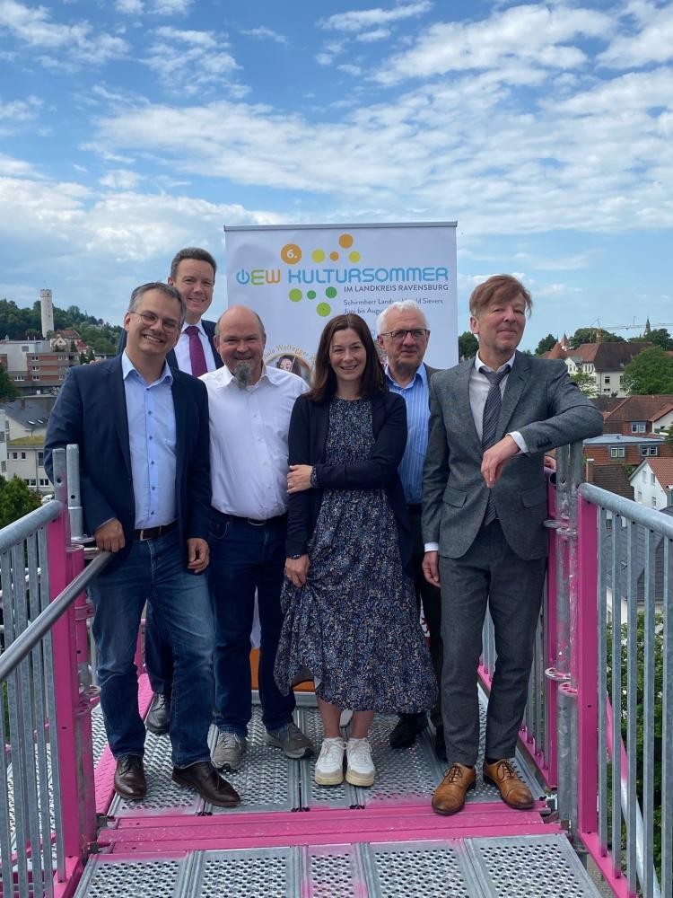 Auf einem Gerüst stehen mehrere gehoben gekleidete Menschen verschiedenen Geschlechts. Sie stehen nebeneinander und lächeln in die Richtung des/der Bildbetrachtenden. Im Hintergrund steht ein Aufsteller mit der Aufschrift OEW-Kultursommer. Es herrscht sonniges Wetter.