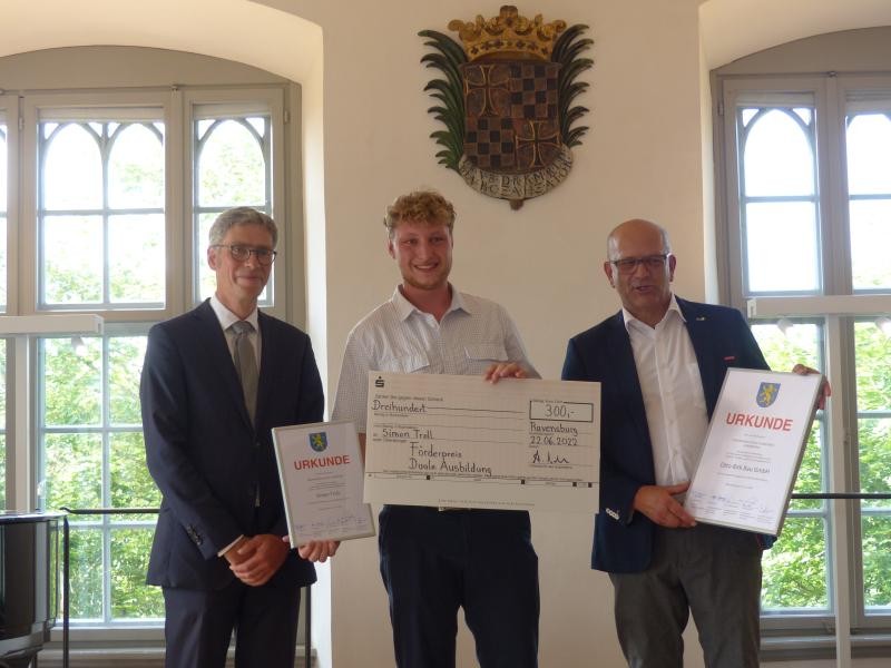 Drei männliche Personen nebneinanderstehend in einem historischen Saal mit einem Wappen an der Wand, unterhalb dessen sie stehen. Sie halten Urkundenpapiere in die Blickperspektive.