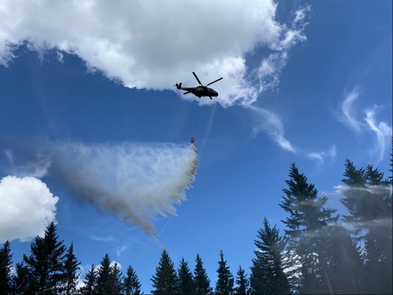 Hubschrauber wirft Löschwasser ab Ein Hubschrauber wirft Löschwasser über einem Wald ab. Es herrscht sommerliches Wetter mit einzelnen Quellwolken am Himmel.