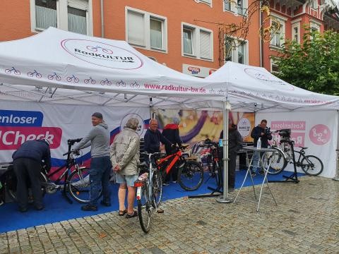 Unter zwei aufgebauten weißen Zelten in einer Innenstadt prüfen und reparieren einige Männer verschiedene Fahrräder. Die Fahrradbesitzenden stehen jeweils daneben, sehen zu, interagieren mit den Mechanikern und warten.