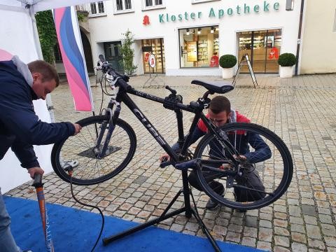 Zwei Männer prüfen und reparieren ein Fahrrad, das auf einem Stativ steht, in einer Fußgängerzone in einer Innenstadt. Im Hintergrund ist eine Apotheke zu sehen.