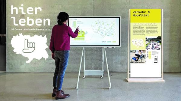 interaktive Ausstellung zu 50 Jahren Landkreis Ravensburg In einem Raum eine Betonwand mit der weißen Aufschrift "hier leben - 50 Jahre Landkreis Ravensburg" mit einer Darstellung des Kreisgebiets, weiß ausgefüllt mit einer aufzeigenden Hand als Icon im Zentrum. Vor der Wand und rechts von der weißen Abbildung eine weibliche Person, die einer interaktiven digitalen Bildschirm-Schautafel zugewandt ist und mit ihrem Finger etwas anwählt. Rechts neben dem aufgestellten Bildschirm eine bedruckte Schautafel an einem Holzaufsteller. Die Überschrift der Tafel lautet: Verkehr und Mobilität.