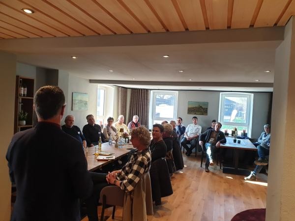 in einem großen Raum sitzen mehrere Personen an Tischen und hören einer ihnen zugewandten und von der Rückseite zu erkennenden männlichen stehenden Person zu.