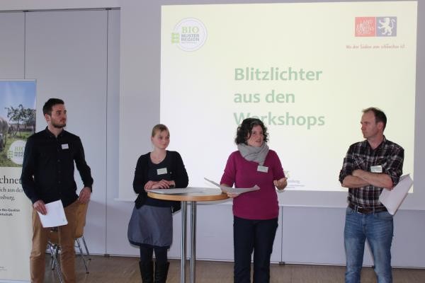 Vier Personen, männlich und weiblich stehen vor einer Vortragsleinwand, auf der geschrieben zu sehen ist: Blitzlichter aus den Workshops