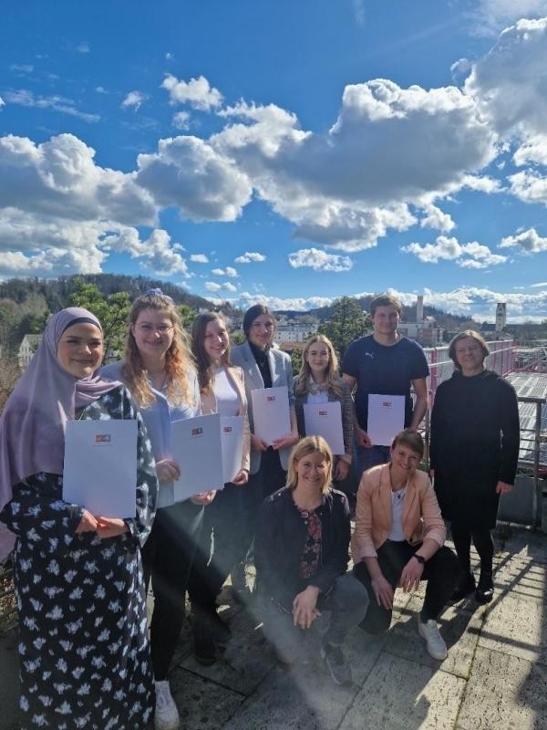 Abschlussfeier der Auszubildenden mehrere junge männliche und weibliche Menschen halten ein Dokument in den Händen und stehen aufgereiht nebeneinander auf einem Terrassendach. Die Ausbildungsleiterinnen knien vor den stehenden Personen.