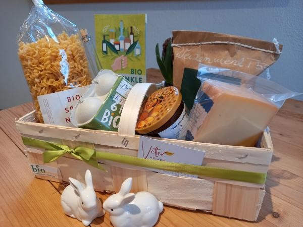 ein quaderförmiger Flechtholzkorb mit verschiedenen Lebensmitteln wie Nudeln, Käse, Honig, Eier, mit österlich grünem Band dekoriert.
