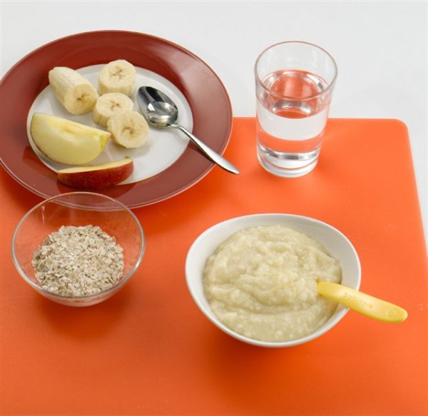 Babys erster Brei auf einem Tisch eine Schüssel mit beige-gelbem Brei, eine kleinere Schüssel mit Haferflocken, ein Teller mit Bananenstücken und Apfelstücken und einem Löffel. Daneben ein Glas mit klarem Wasser.