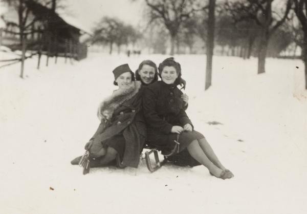 Winter in den 1930ern. auf einer Schwarz-weiß-Fotografie drei jugendliche Mädchen in Winterkleidung auf einem schneebedeckten Weg auf einem Holzschlitten sitzend