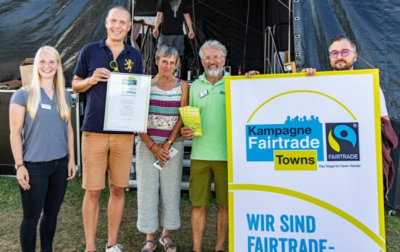 Die Steuerungsgruppe des Fairtrade-Landkreises freut sich über die Rezertifizierung Fünf Personen nebeneinander, die in die Kamera schauen und die Urkunde halten und ein Plakat