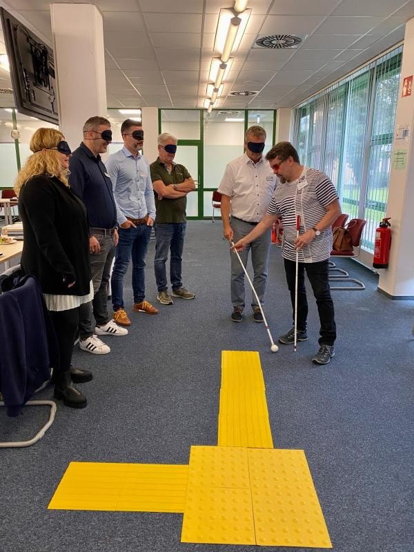 Alltag aus der Perspektive von Menschen mit Behinderungen nachempfinden 6 Menschen stehen in einem Raum mit Augenmasken umgebunden. Zwei davon halten einen Blindenstock in der Hand. Vor diesen Personen liegt eine gelbe Blindenführungswegmarkierung.