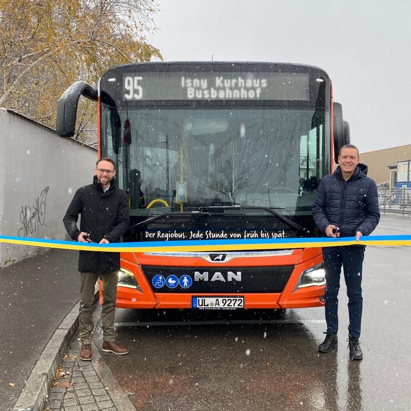 Zwei Männer stehen vor dem neuen Schnellbus und durchschneiden ein Band zur Einweihung der neuen Buslinie