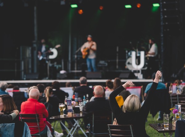 vor einer Open-Air-Bühne sitzen Menschen, von denen eine Person die Hände zum Jubel erhoben hat. Auf der Bühne spielt eine Musikband.