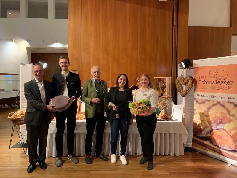 Käserei Bauhofer Bodnegg, Brauerei Clemens Härle Leutkirch, Schmalegger Beck Schmalegg Fünf Personen stehen nebeneinander in einer Reihe mit Blick Richtung Kamera. Darunter drei Männer und zwei Frauen. Sie platzieren in ihren Händen jeweils Brotgebäck, Bierflaschen oder einen Käselaib.