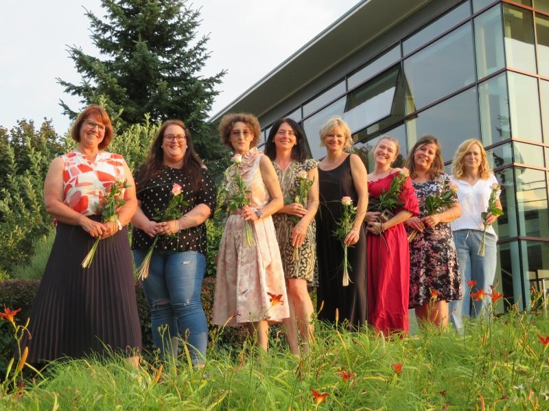 8 Frauen stehen nebeneinander vor einem Glasgebäude und halten jeweils eine Rose.