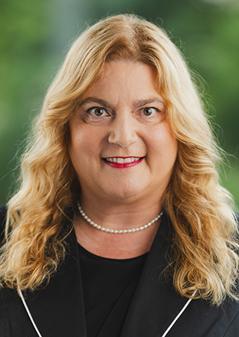 Eine Frau mittleren Alters mit blondem langem gescheiteltem Haar, mit Halskette und dunklem Jackett mit schwarzem Oberteil darunter