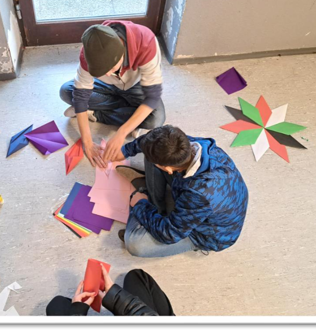 Mann und Kind sitzen nebeneinander auf dem Boden und basteln gemeinsam au sbunten Papieren einen Stern