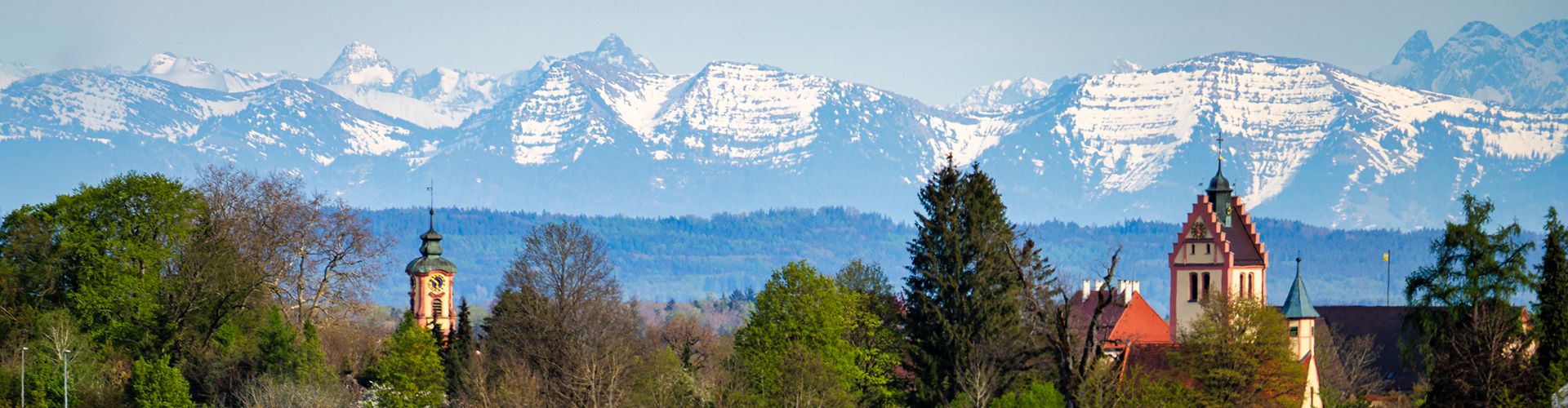 Eine Weitwinkelaufnahme mit dem Blick auf Kisslegg, umrahmt von Wiesen, Bäumen und Waldstücken, im Hintergrund die Alpenbergkette bei lichtem Wetter in einer Frühlingsstimmung.