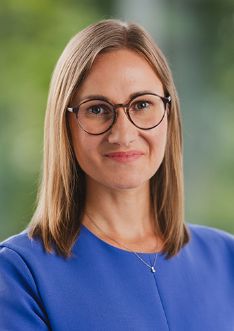 braunhaarige Frau mit schulterlangem glattem Haar und Brille im Alter zwischen 30 und 40 Jahren, die in die Betrachterperspektive schaut und lächelt.