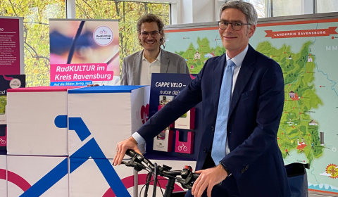 Eröffnung RadGalerie Zwei Männer stehen vor der Kulisse der Ausstellung und blicken in die Kamera. Ein Mann davon steht an einem Fahrrad.