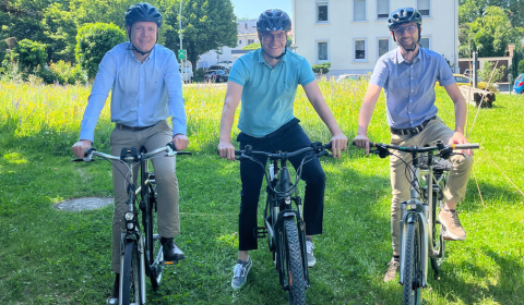 Auf einer Wiese sitzen drei Männer nebeneinander auf ihrem Fahrrad mit Helm