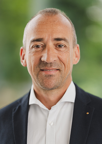 Mann mittleren Alters mit hoher Stirn, ohne Bart und dunklen Augenbrauen. Er trägt ein dunkles Jackett und ein weißes Hemd